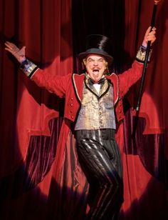 a man in a red jacket and top hat with his hands up while standing on stage