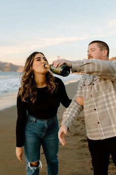 a man and woman standing on top of a beach next to the ocean drinking from bottles