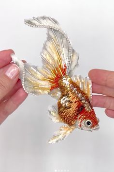 a hand holding a goldfish ornament in it's right hand and another hand is holding the fish