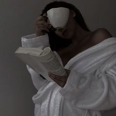 a woman in a bathrobe reading a book and holding a cup to her face