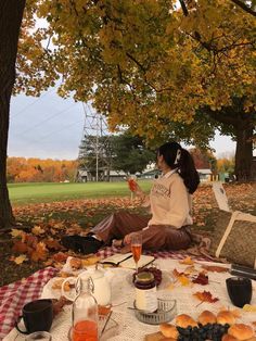 a woman sitting at a picnic table with food and drinks in front of her on an autumn day