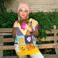 a woman sitting on a bench holding a pink cup in her right hand and wearing a multicolored sweater