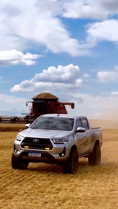 a silver truck driving down a dirt road next to a large red combine behind it