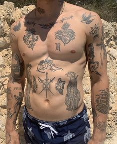 a man with many tattoos on his chest standing next to a rock wall and looking at the camera