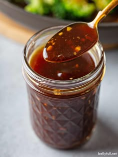 a spoon full of bbq sauce on top of a plate with broccoli in the background