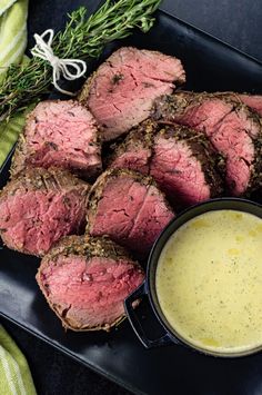 some meat is on a black plate next to a bowl of sauce and green sprigs