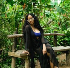 a woman sitting on top of a wooden bench in front of green plants and trees