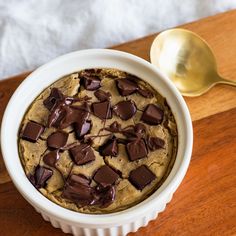 a chocolate chip cookie in a white bowl next to a spoon on a wooden table