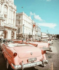 an old pink car parked in front of a white building on a city street with tall buildings behind it