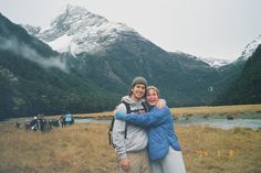 two people hugging each other in the middle of a field with mountains in the background