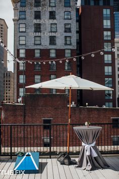 an umbrella and table set up on a deck in front of tall buildings with string lights