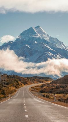 an empty road in front of a large mountain with snow on the top and clouds flying over it