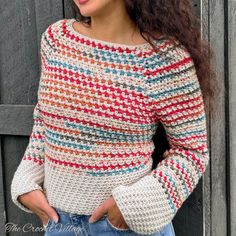 a woman standing in front of a wooden fence wearing a colorful sweater and denim jeans