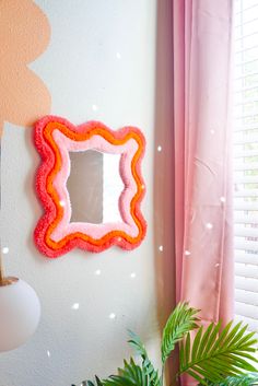 a pink and orange mirror on the wall next to a potted plant in front of a window