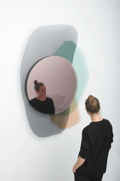 a woman standing in front of a white wall with a circular mirror hanging on it's side