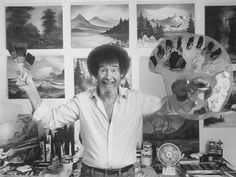 a man standing in front of many paintings and holding a paintbrush up to his face