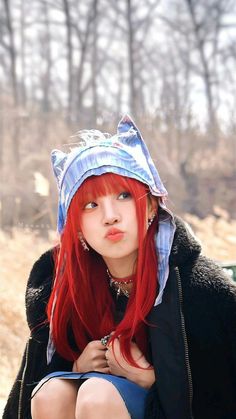 a young woman with red hair wearing a blue hat and black jacket sitting on the ground