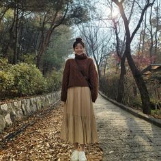 a woman standing in the middle of a leaf covered path wearing a brown sweater and beige pleated skirt