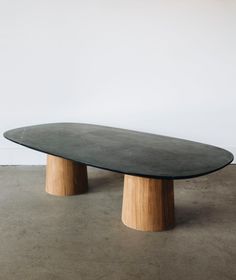 an oval table with two wooden legs and a black top on concrete flooring in front of a white wall