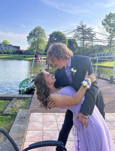 a man in a tuxedo is hugging a young woman on the dock near a lake