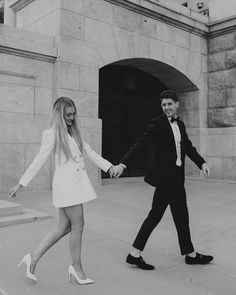 a man and woman holding hands while walking down the street in front of a building