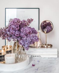 purple flowers in a glass vase on a marble counter top next to books and candles