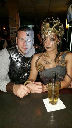 two people sitting at a table with drinks in front of their faces, one wearing a mask and the other holding a drink