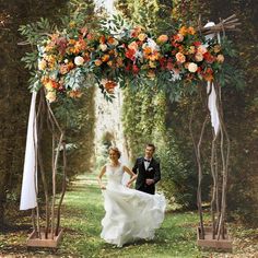 a bride and groom walking under an arch decorated with orange, yellow and white flowers