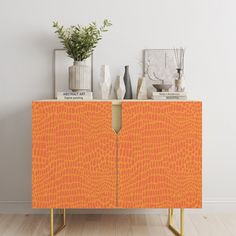 an orange cabinet with gold legs in front of a white wall and vases on top