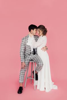 a man and woman sitting on top of a stool in front of a pink background