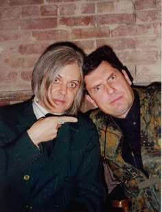 two people sitting next to each other in front of a brick wall and pointing at the camera