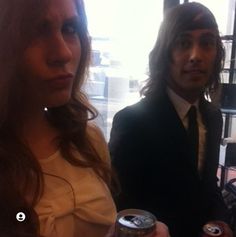 two people standing next to each other in front of a window holding cans of beer