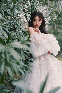 a woman in a white dress standing next to some trees and bushes with her hands on her chest