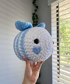 a hand holding up a crocheted blue and white stuffed animal in front of a window