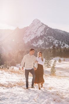 a man and woman walking through the snow in front of a mountain with their arms around each other