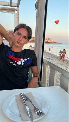 a man sitting at a table with a plate and fork in front of him on the beach