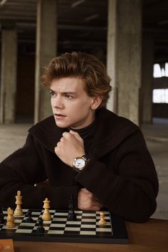 a young man sitting at a chess board with his hand on his chin and looking to the side