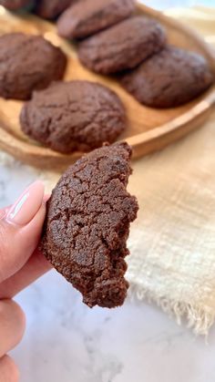 a hand holding a chocolate cookie in front of some cookies on a wooden plate and napkin