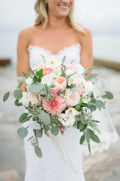 a woman holding a bouquet of flowers and greenery in front of the ocean on her wedding day