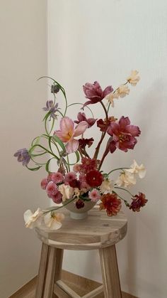 an arrangement of flowers in a vase sitting on a small wooden stool next to a white wall