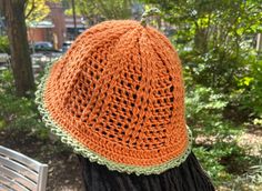 an orange knitted hat sitting on top of a wooden bench next to trees and bushes