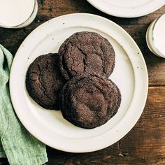 three chocolate cookies on a white plate next to two glasses of milk and a green napkin