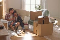 a man and woman sitting on a couch surrounded by boxes in a room with windows