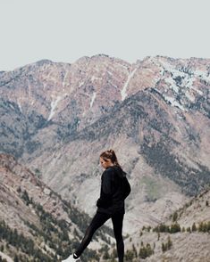 a woman standing on top of a mountain with her feet in the air and mountains behind her