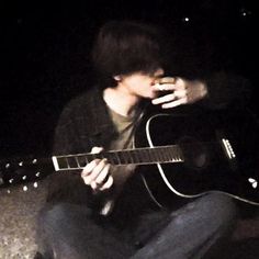 a man sitting on the ground playing an acoustic guitar at night with his mouth open