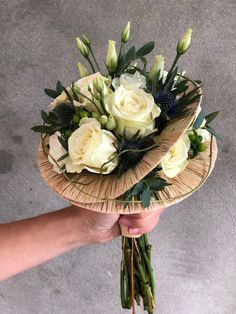 a hand holding a bouquet of white roses and greenery in a straw hat on the ground