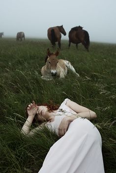 a woman laying in the grass with horses behind her