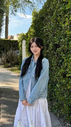 a woman standing next to a hedge holding a white purse and wearing a denim jacket