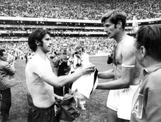 two men shaking hands in front of an audience at a sporting event, one holding a towel and the other wearing shorts