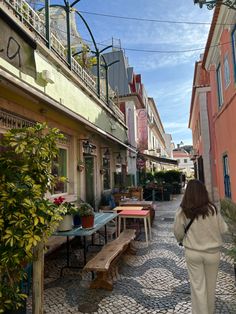 a woman walking down a cobblestone street next to tables and chairs with plants on them